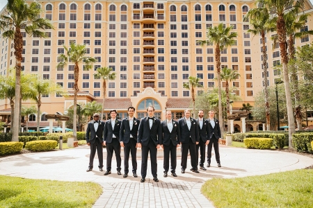 8 men in matching black tuxedos stand outdoors in a courtyard surrounded by palm trees with a towering hotel behind them. Rosen Shingle Creek is perfect for an elegant hotel wedding.