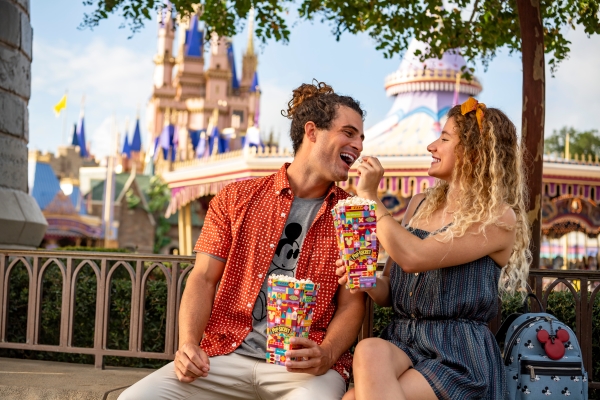 A man and a woman sit on a bench feeding each other popcorn with a carousel and Cinderella Castle in the background.