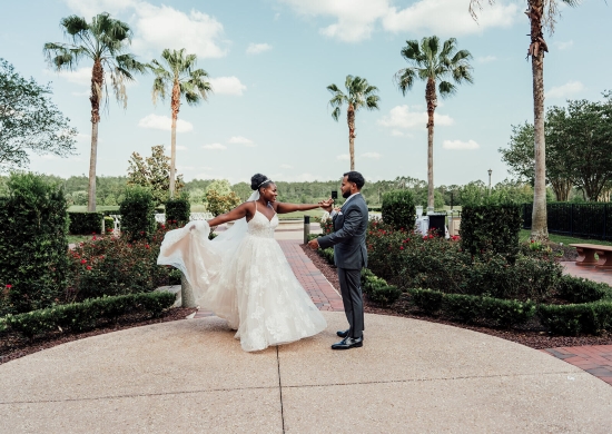 A woman in a long white gown holds her dress as she is spun by a man in a gray suit in a courtyard surrounded by roses and palm trees.