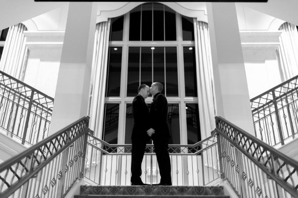 A black and white image of two grooms on a stairway landing in front of a towering window. 