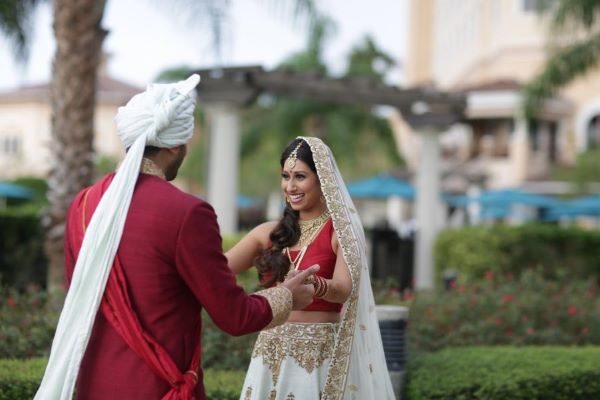 A bride in a red and white sari happily greets a groom in a red coat with a long white turban outdoors, a romantic first look from Rosen Shingle Creek.