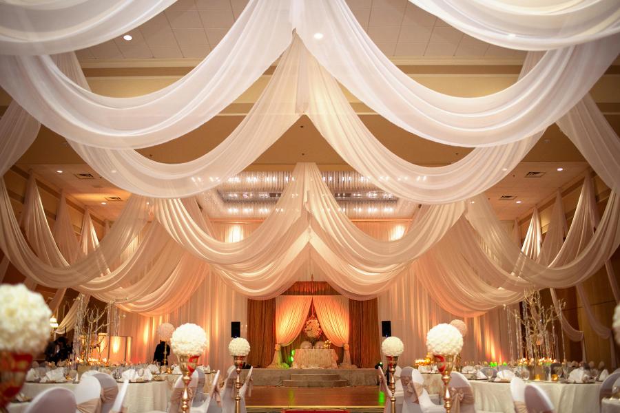 A beautiful ballroom with drapery hanging from the ceiling, amber glowing lights, and tall floral centerpieces on round tables. 