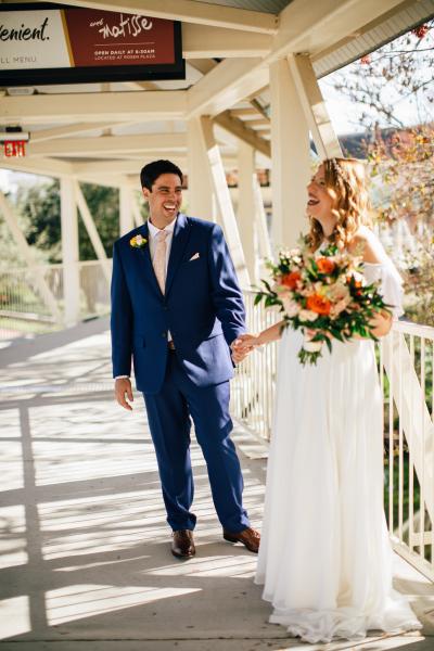 A bride holding a bouquet and a groom in a blue suit stand on a covered breezeway while laughing, a first look from a beautiful wedding at Rosen Plaza. 