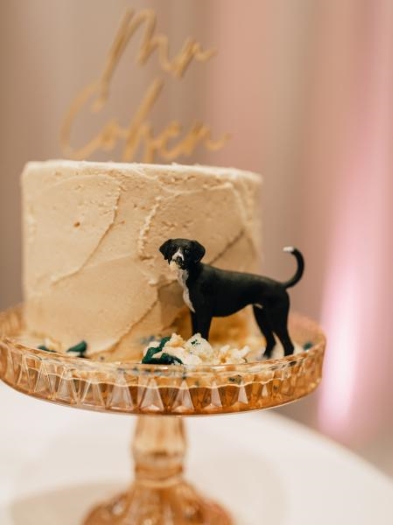 A small, one-tier cake on a crystal cake stand adorned with a black and white dog figurine posed to look like it's eating the cake. A cake topper is a cute way to incorporate your pet in your wedding.