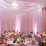 An event setting with sheer white drapery hanging from the ceilings and walls, pale pink uplighting, and round tables set with candles and florals. A hotel wedding can be a glamorous event in an elegant ballroom space.