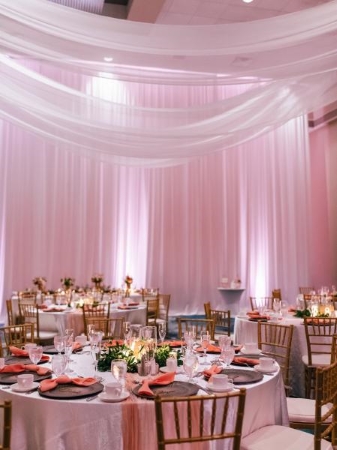 An event setting with sheer white drapery hanging from the ceilings and walls, pale pink uplighting, and round tables set with candles and florals. A hotel wedding can be a glamorous event in an elegant ballroom space.