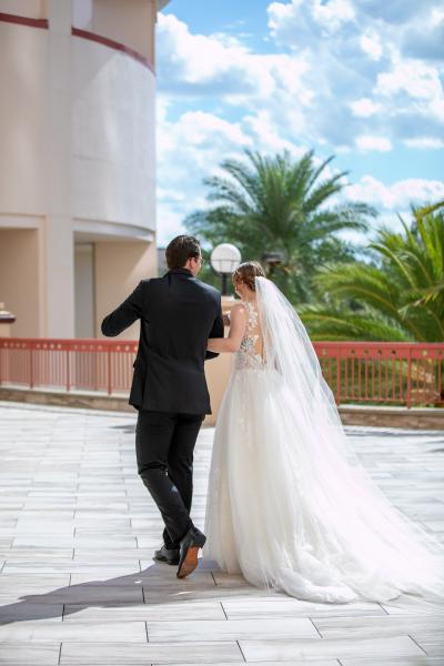 A bride and groom on an outdoor terrace overlooking blue sky and the tops of green palm trees walk away from the camera after a private first look moment.