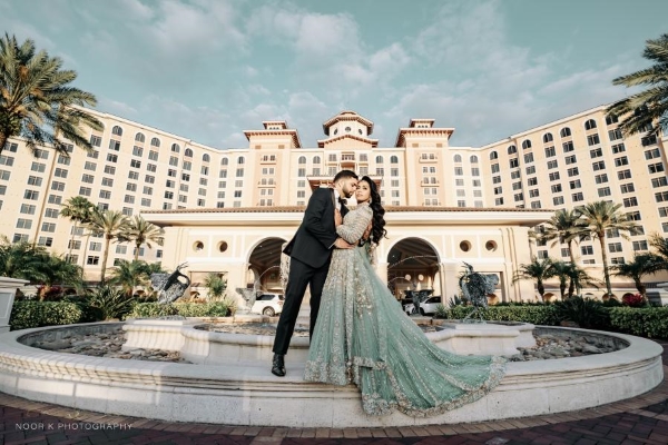 A bride in an ornate blue gown embraces her groom standing on the ledge of a fountain in front of a sprawling hotel. 