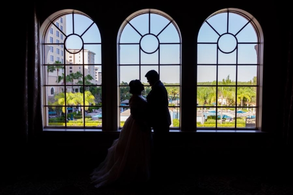 A couple silhouetted in front of three tall arched windows overlooking swimming pools and palm trees in the distance.