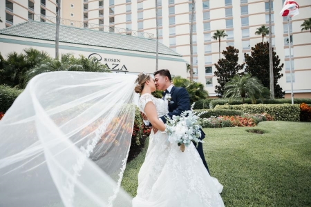 A bride and groom stand on a green lawn with the entrance to Rosen Plaza behind them. Rosen Plaza is an elegant destination for a hotel wedding.