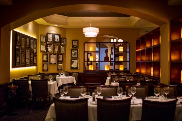 A dimly lit restaurant with leather chairs and white linen tablecloths. The walls are decorated in amber glowing lights and dozens of framed caricatures. 