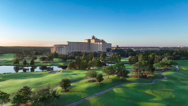 An aerial shot of a tall hotel amidst green grass, trees, and a serene lake. 