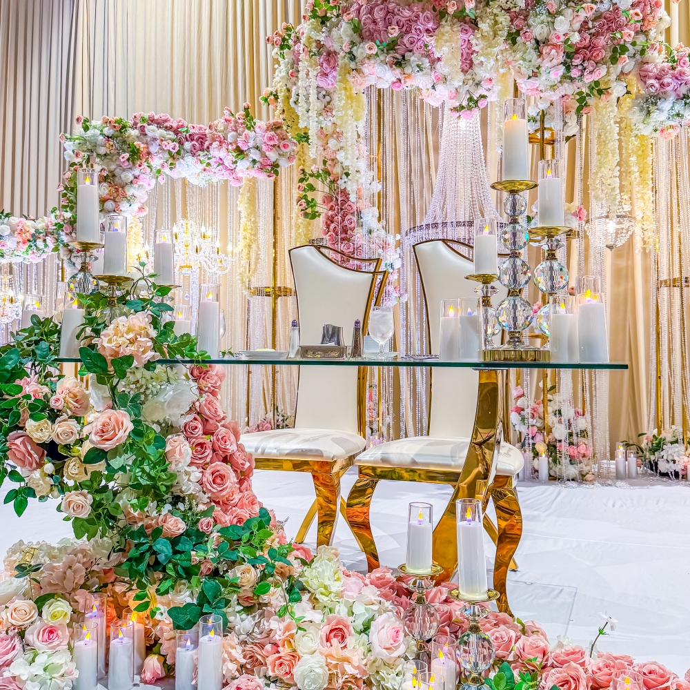 A lavish sweetheart table setup with a glass table, arches of flowers, and two white and gold chairs beside a cascade of flowers and candles. 