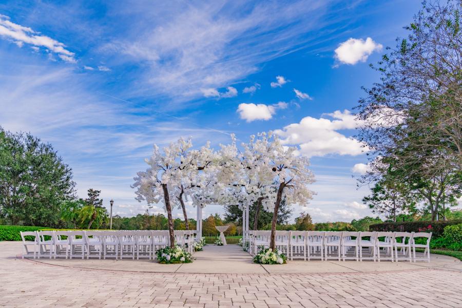 Outdoors on a patio, rows of white chairs flank several large arches of trees with white blossoms.