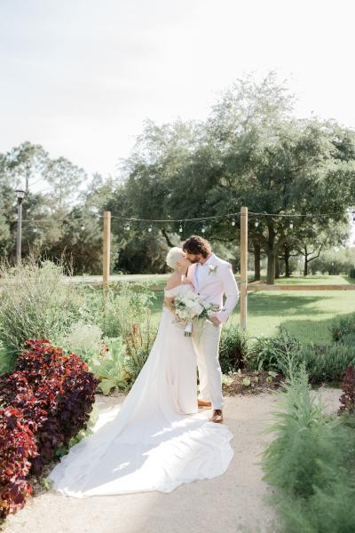 A bride and a groom in a light pink suit kiss in an elegant garden setting with a row of string lights behind them. 