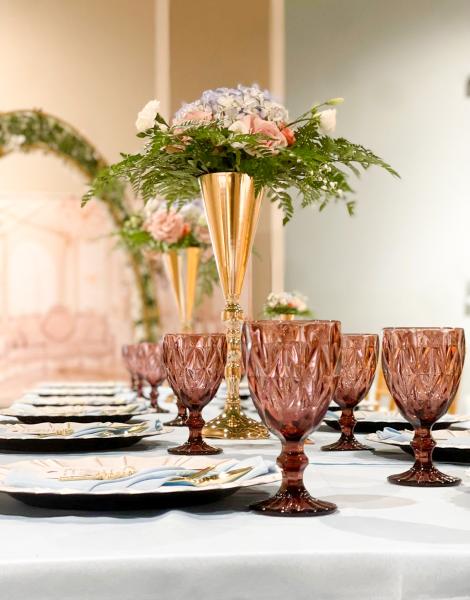 A close-up of a table setting with rose colored goblets and gold vases with greenery and flowers. 