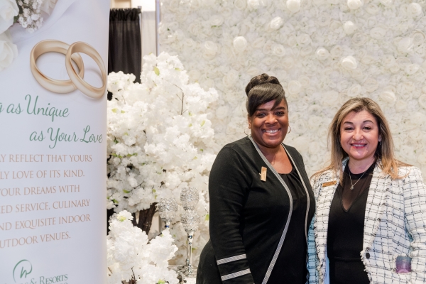 Two women in professional attire standing in front of a white floral expo backdrop. Meet the Rosen Weddings team at the Wedding Expo!