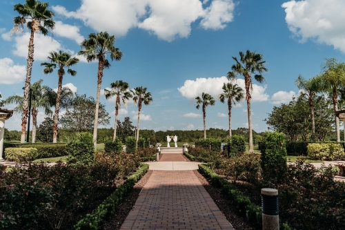 A cobblestone path leading to a distant arch draped with white fabric, surrounded by tall palm trees and set against a blue, cloudy sky. Hotel weddings can take advantage of beautiful landscaping.