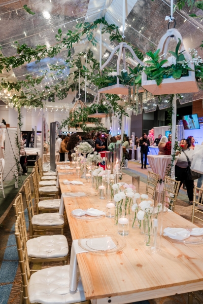 A sample table setting at a wedding expo with a long wooden table, gold chiavari chairs, simple white florals, and greenery hanging from a transparent tent overhead. 