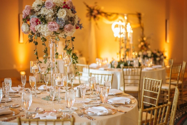 A wedding table setting with gold chiavari chairs, tall centerpieces, and white linens. A yellow neon sign glows in the background out of focus. Let Rosen Weddings help you customize your wedding menu.