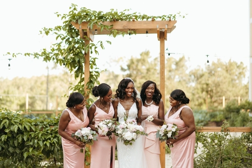 A bride in a white gown and four bridesmaids in light pink gowns hold bouquets and stand beneath a wooden archway covered in vines.