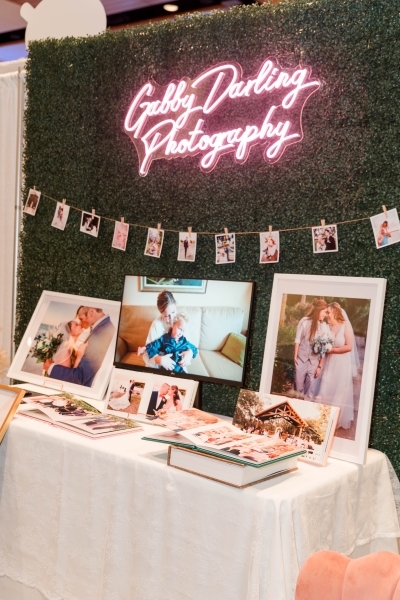 A wedding expo photography vendor with a neon sign, hanging photos, and a table of photo albums and framed pictures. 