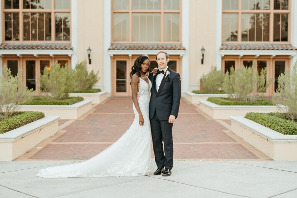 A woman in a long, white dress and a man in a black tuxedo stand in front of a pathway lined with raised landscaping and leading to multi-story glass windows. 