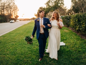 A bride wearing an A-line wedding dress holding a glass of champagne and walking in arms in the grass with her groom.