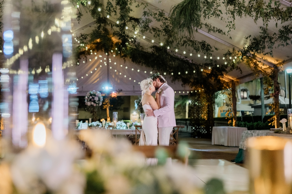 A bride and groom dance alone on the middle of a dance floor in a tent lined in greenery and bistro lights. 