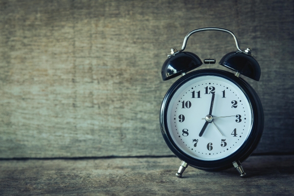 A black alarm clock with a white face sitting on a wooden surface against a wooden background. 