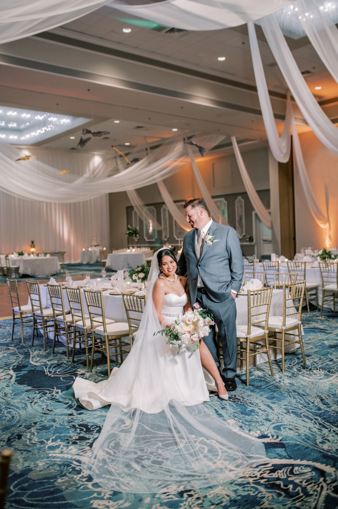 A bride sits in a chair in a formal ballroom with blue carpet and drapery hanging from the ceiling, her groom standing beside her. 