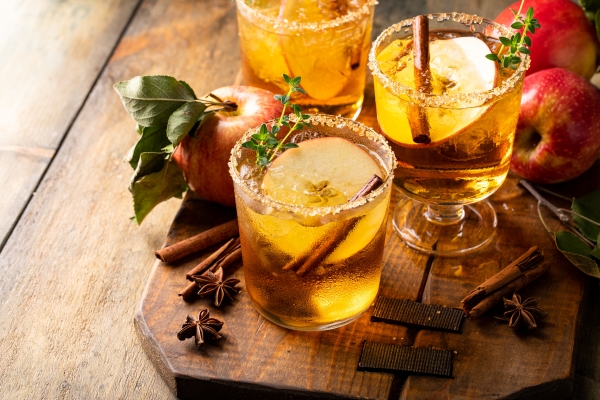Three glasses garnished with thyme, apple slices, and cinnamon sticks surrounded by spices and fresh apples, creating the perfect fall wedding cocktail.
