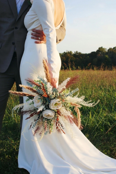 A woman in a long sleeved white dress holding a large autumnal bouquet embraced by a man in a gray suit standing in a grassy field.