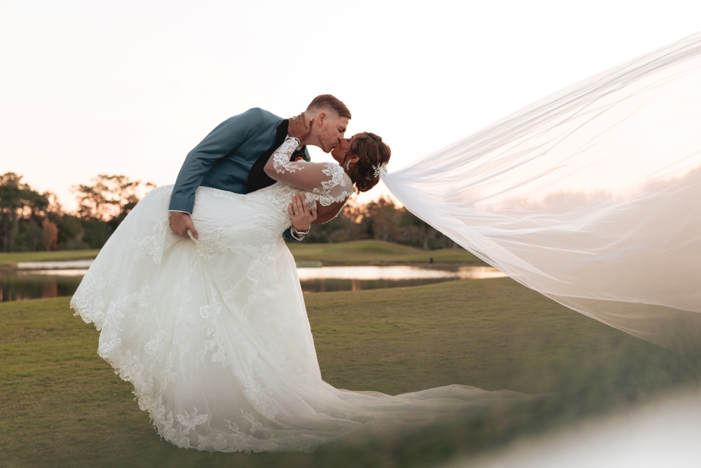 A groom dips his bride as he kisses her in a twilight outdoor setting, her veil blowing in the breeze.