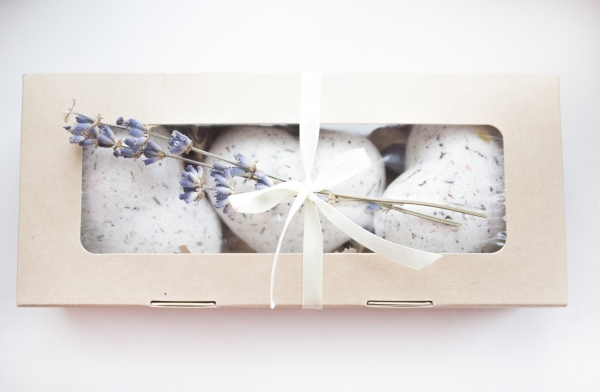 A paper box with a see-through top showing multiple white bath bombs inside, tied with white ribbon and a sprig of lavender.
