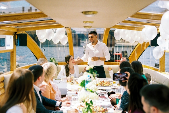A group of people dining on a boat surrounded by white balloons while a man in a white shirt delivers a speech.