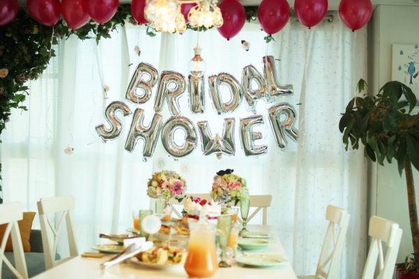 A white table set with flowers and brunch dishes in front of a window with sheer curtains and silver balloons that read Bridal Shower.