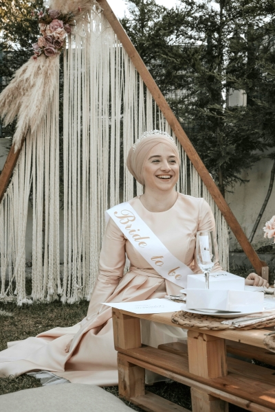 A woman in a pink dress and head wrap smiles wearing a Bride to Be sash seated on the lawn in front of a triangular arch with flowers and hanging rope.