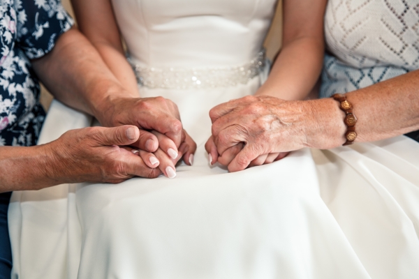A closeup of the lap of woman in a bridal gown holding hands with two older women on either side.