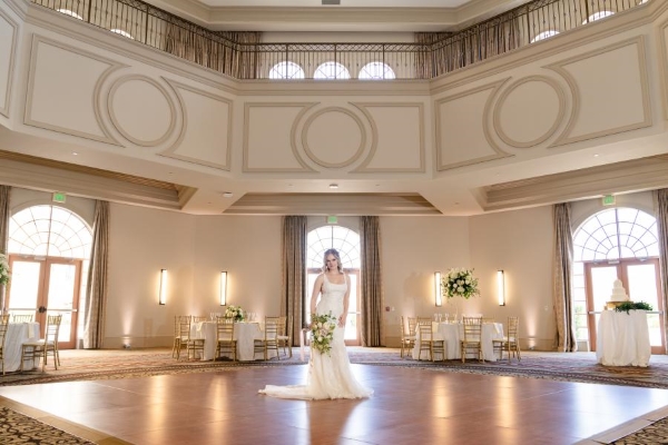 A woman in a white dress holding a bouquet stands in the middle of a dance floor in a two-story ballroom with a surrounding balcony and large windows behind her.