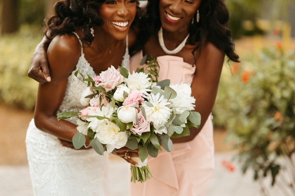 A woman in a bridal gown and a woman in a pink bridesmaid dress hold a pink and white bouquet, bent over with laughter.
