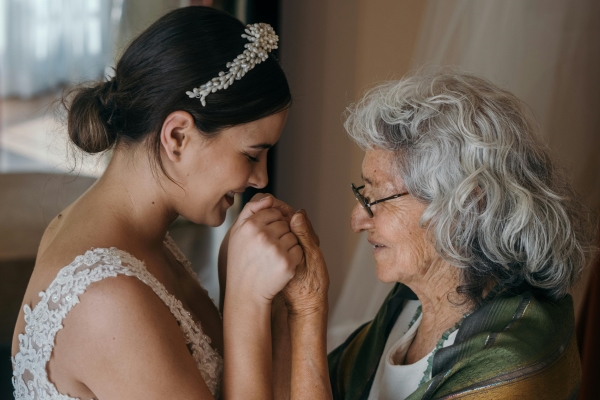 A woman in a white dress and pearl headband holds hands with an older woman wearing glasses and a green shawl.