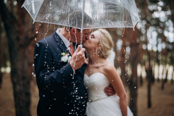A bride and groom kiss outdoors in the rain beneath a clear open umbrella.