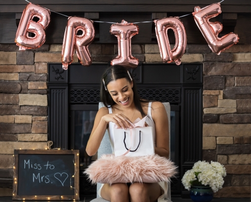 A woman in a white dress sits in front of a stone fireplace beneath pink balloons that read "bride." She sits next to a sign that reads "Miss to Mrs." and opens a gift bag.