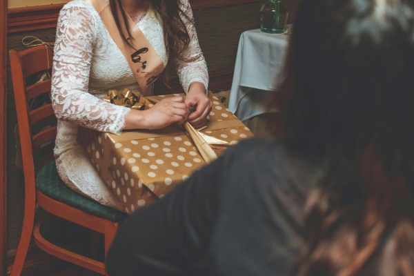 The torso of a woman in a lacy white dress opening a gift wrapped in gold and white polka dotted wrapping paper.