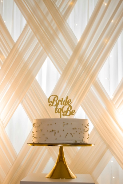 A white cake with a gold topper that reads Bride to Be in front of latticed cream colored curtains.