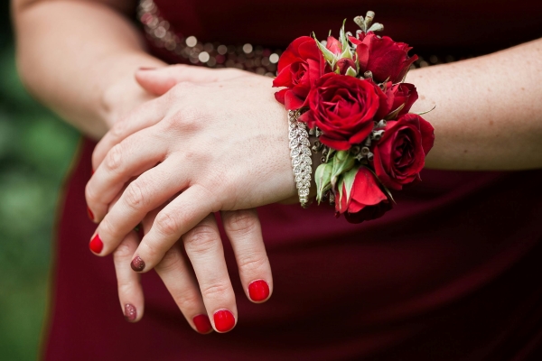 A closeup of the hands of a woman in a maroon dress with red nail polish and a corsage made of red roses.
