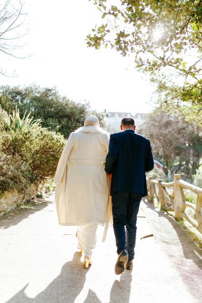 A bride in a coat walks beside a man in a dark suit along a path surrounded in greenery.