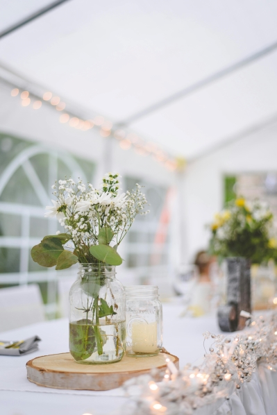 In a large white tent, mason jars filled with simple greenery and a white candle sit on log slice centerpieces on a white table.