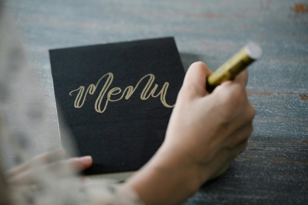 A close-up of a hand with a gold pen writing "menu" on a black piece of paper in calligraphy.